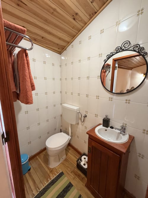 Banheiro rústico na villa Casa da Capela com teto de vigas de madeira, toalhas de terracota, pia de coluna e paredes com azulejos creme