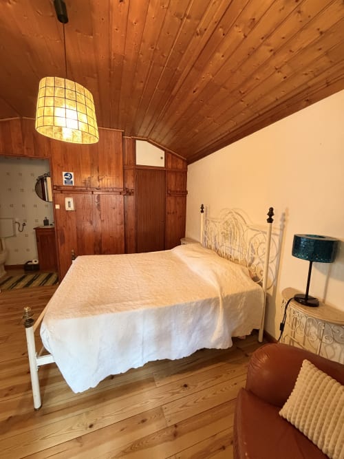 Quarto aconchegante na villa Casas do Sinhel com cama de ferro branco, vigas de madeira, luminária pendente e charme rústico em Roda Fundeira