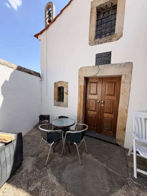 Pátio rústico da Casas do Sinhel com mesa de refeições, cadeiras, entrada da capela de pedra e vistas de montanha