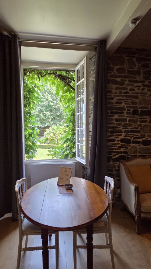 Salle à manger avec table ronde en bois au Manoir de la Bégaudière, Mont-Dol, avec vue sur le jardin verdoyant par la fenêtre ouverte