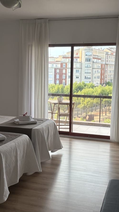 Dormitorio luminoso en Muro 13 El Mirador del Espolón con camas, puertas abiertas a la terraza, cortinas blancas y vistas a la ciudad de Logroño