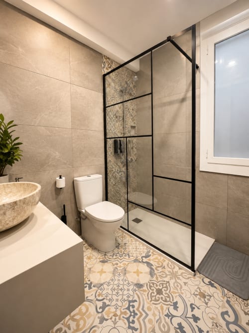 Baño moderno con azulejos beige, ducha con marco negro, inodoro blanco y suelo con patrón decorativo en Muro