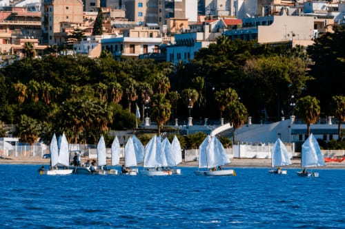 Barche a vela bianche su acque azzurre a Reggio Calabria con edifici colorati e palme che costeggiano la riva