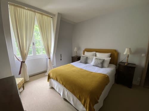 Bright bedroom with yellow bed throw, mustard headboard, cream curtains, and forest view at Domaine de Bize Mirepoix