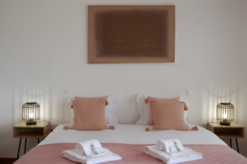 Quarto elegante em Be SilverCoast Serra D'El Rei com almofadas em tons de rosa, roupas de cama brancas, luminárias laterais e arte abstrata na parede