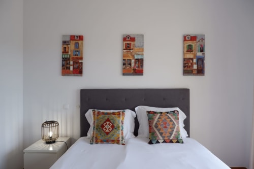Quarto com cabeceira escura, roupa de cama branca, almofadas com padrões coloridos e três pequenas pinturas de casarões acima chez Be