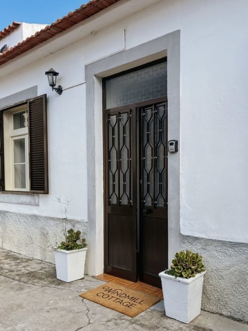 Entrada da Windmill Cottage nas Villas Simone Porto Santo com portas pretas ornamentadas, plantas em vasos brancos e tapete de boas-vindas