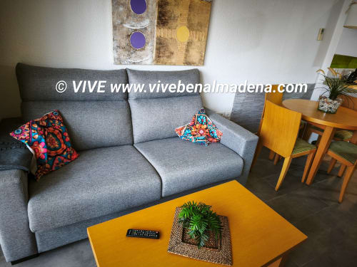 Gray sofa with colorful pillows, yellow table with plant, wooden dining set, and wall art in VIVE Benalmádena apartment