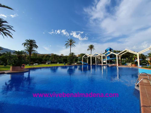 Bright blue resort pool with white cabanas, palm trees, and green gardens at VIVE Benalmádena beachfront hotel under sunny
