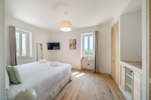 Bright modern bedroom in Grasse villa with white bed, wooden flooring, flat-screen TV, and natural light from windows