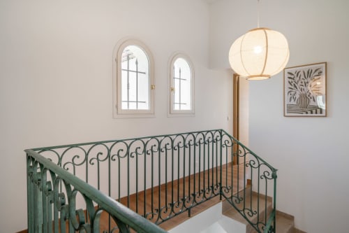 Elegant staircase landing in Grasse villa with wrought iron railing, arched windows, pendant lamp, and botanical artwork