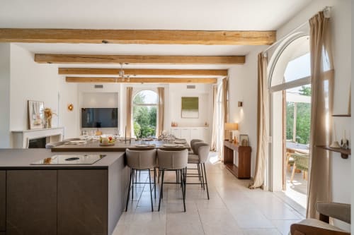 Modern villa dining and living area with exposed beams, kitchen island, and French doors opening to terrace at M Résidences,