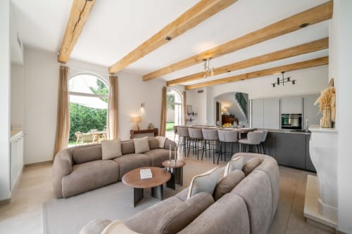 Modern villa living room with exposed wooden beams, curved gray sofas, open kitchen, and arched doors to terrace in Grasse