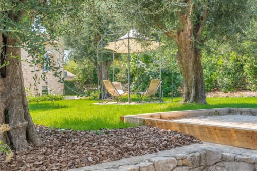 Sunny villa garden with lounging area under umbrella, mature trees, sandy sandbox, and stone terrace at M Résidences, Grasse