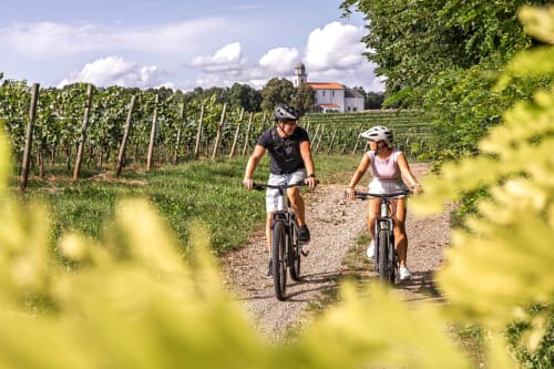 Deux cyclistes roulent sur un chemin de gravel à travers les vignobles près de Mons-Boubert, avec une église au loin sous un ciel bleu.