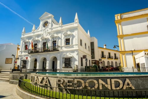 Edificio colonial blanco del Hotel Plaza Chica con torre de reloj en Cartaya, España, con fuente y cielo azul