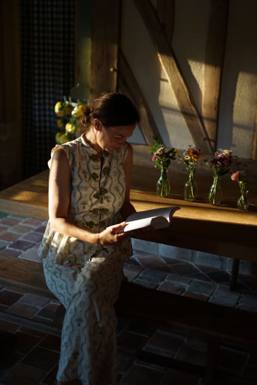 Femme lisant près d'une fenêtre ensoleillée à la Ferme de La Roquerie, avec des fleurs fraîches dans des vases en verre sur une table en bois