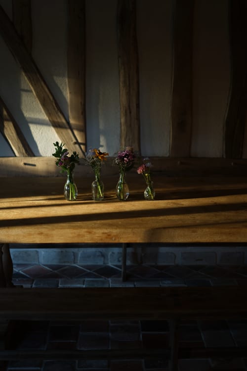 Quatre petits vases en verre avec des fleurs colorées sur la cheminée de la Ferme de la Roquerie, baignés de lumière avec des rideaux drapés