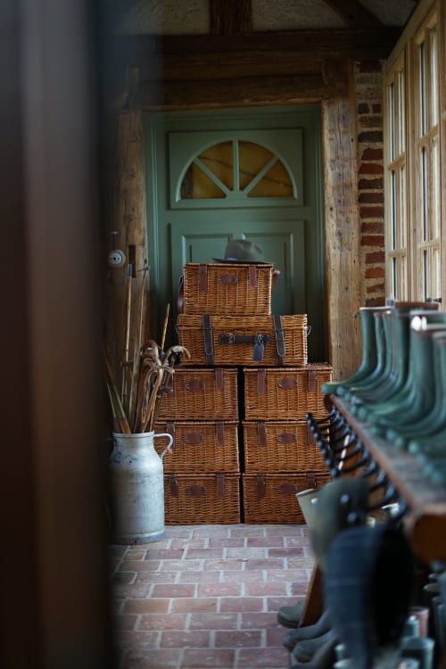 Entrée rustique de la Maison de la Roquerie avec valises en osier empilées, porte verte et sol en brique