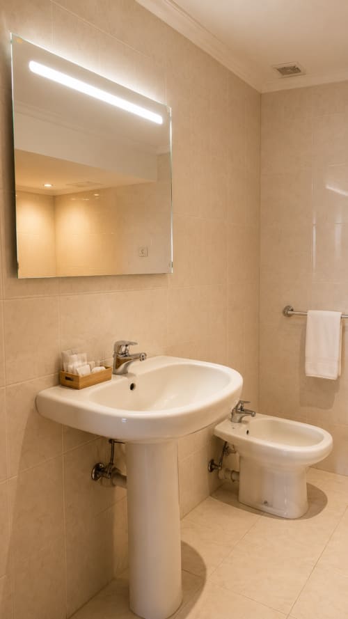 Baño moderno en habitación del hotel Matzuri con dos lavabos de pedestal, espejo iluminado y azulejos beige
