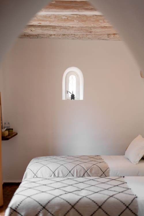 Minimalist bedroom at Domaine Belhazar, Essaouira with arched window, white bedding, exposed wooden ceiling beams, and pale