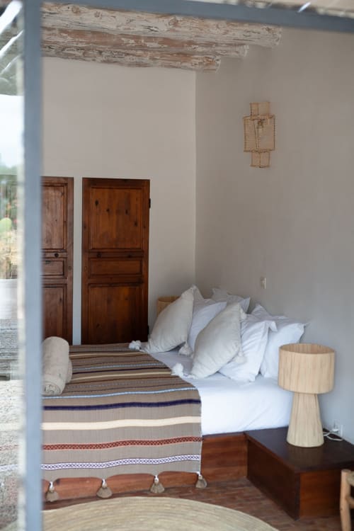 Rustic bedroom at Domaine Belhazar, Essaouira with white pillows, striped blanket, wooden doors, exposed beams, and