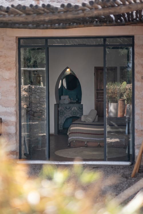 Bedroom at Domaine Belhazar in Essaouira with arched fireplace, striped bed, and potted plants visible through glass doors
