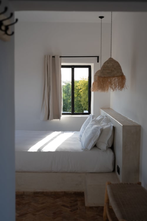 Bright minimalist bedroom at Domaine Belhazar in Essaouira with white bed, woven pendant light, and garden view through