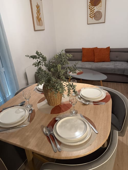 Espace de repas moderne à Appart'NoOm avec table dressée, plante en pot et canapé gris aux coussins orange
