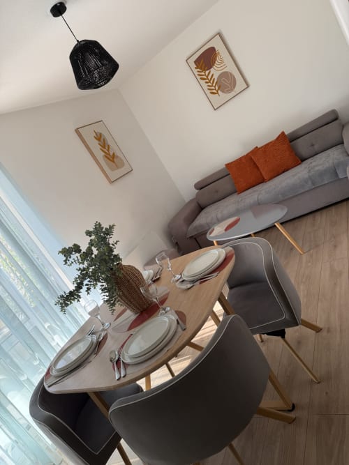 Coin repas chaleureux d'Appart'NoOm avec table dressée, canapé gris, coussins orange et décoration murale botanique