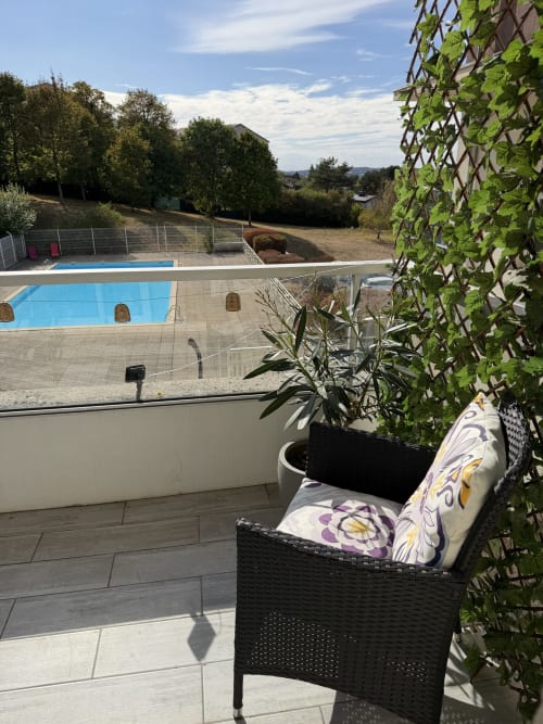 Balcon confortable de l'Appart'NoOm avec vue sur la piscine, chaise en rotin, treillis de lierre et piscine en contrebas