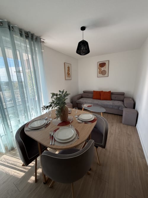 Coin repas cosy avec table dressée, canapé gris, suspension noire et vue sur balcon à Appart'NoOm