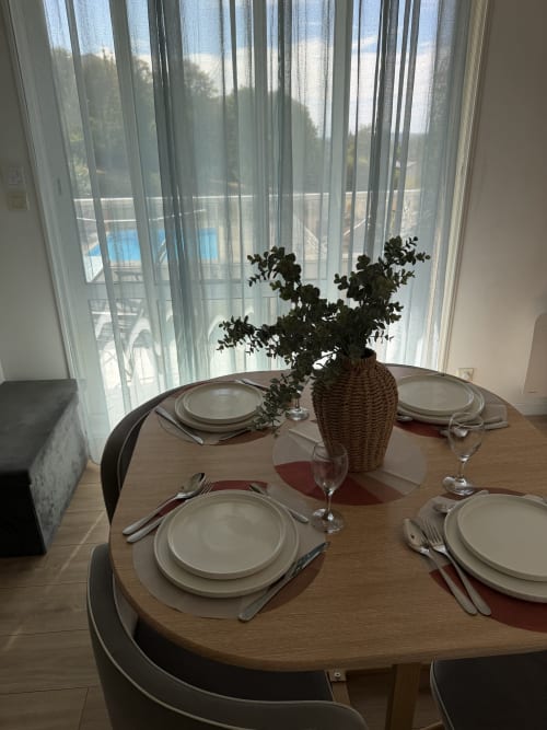 Élégante salle à manger chez Appart'NoOm avec table ronde, couverts, plante en pot et vue sur piscine à travers des rideaux voilages
