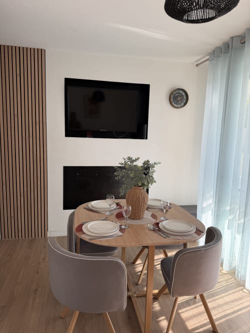 Salle à manger contemporaine chez Appart'NoOm : table ronde en bois, couvert, vase tressé, TV murale, vue sur la piscine