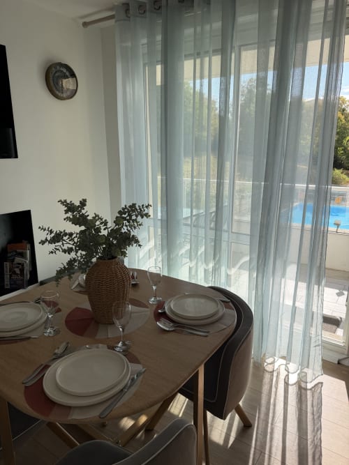 Coin repas cosy à Appart'NoOm avec vue sur la piscine, table dressée, vase en osier et voilages