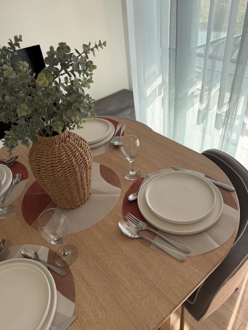 Élégante table de salle à manger à Appart'NoOm avec composition d'eucalyptus, couverts et vue sur la piscine à travers des rideaux voilés