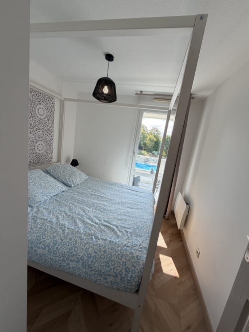 Chambre moderne avec lit à quatre colonnes, luminaire suspendu noir et balcon donnant sur la piscine à Appart'NoOm