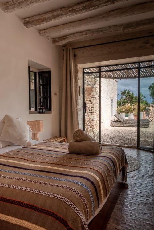 Chambre rustique au Domaine Belhazar avec literie rayée, poutres apparentes, murs en pierre et vue océan depuis la terrasse