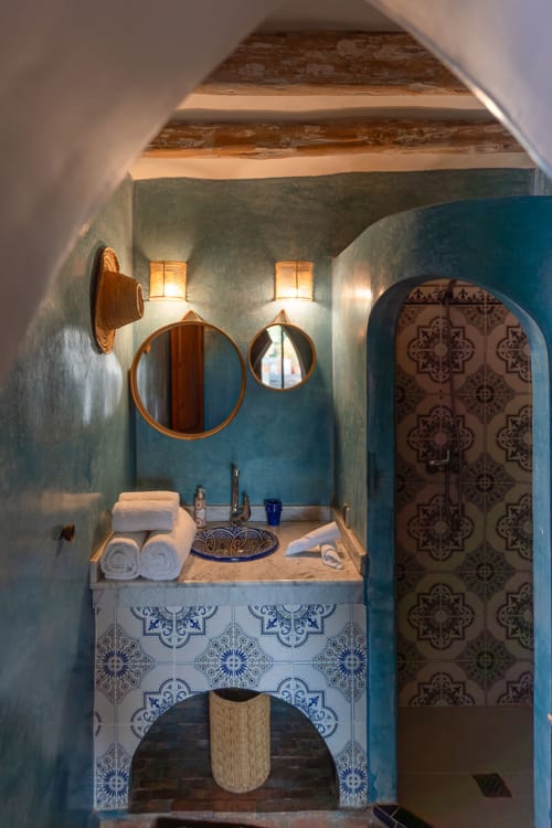 Salle de bains style marocain au Domaine Belhazar avec murs bleus, carrelage orné, miroirs circulaires et luminaires traditionnels.