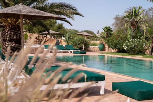 Turquoise pool with white loungers at Domaine Belhazar, Essaouira. Palm trees and stone buildings visible.