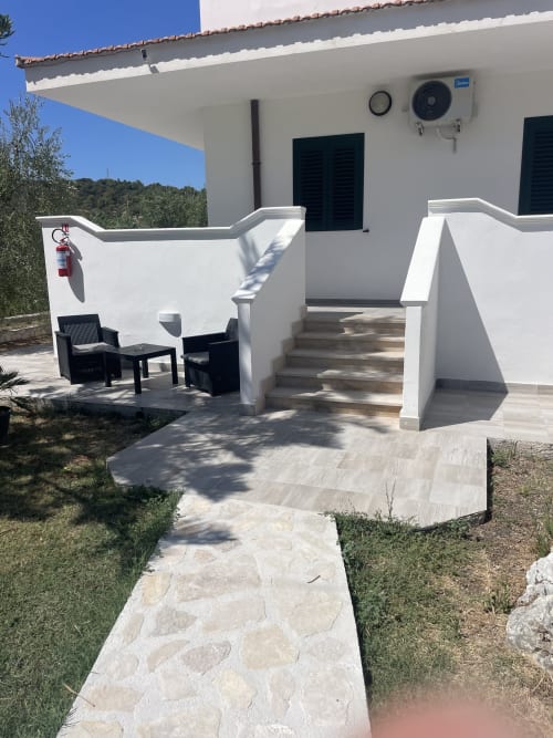 Patio dell'appartamento Villa Angela a Vieste con muri bianchi, zona lounge, sentiero in pietra e vista sul giardino sotto cielo azzurro