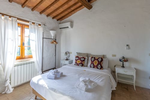 Camera da letto luminosa con soffitto con travi in legno, pareti bianche, letto matrimoniale con cuscini decorativi, finestra con cornice in legno