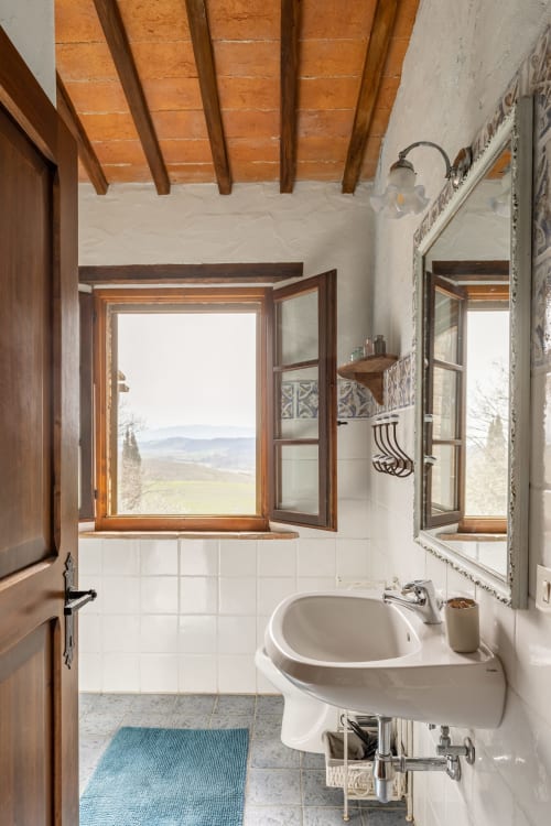 Bagno rustico con travi in legno, piastrelle bianche, lavabo su piedistallo e finestre aperte sulla campagna toscana
