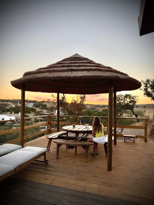 Mulher de camisa amarela jantando sob gazebo coberto de palha na quinta Corvatos Casas do Monte em Almodôvar, Portugal, com vistas do pôr do sol