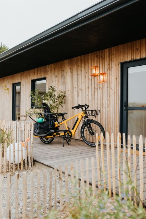 Vélo cargo jaune garé sur la terrasse en bois du gîte Les écuries à Landunvez avec clôture blanche et vue sur le jardin