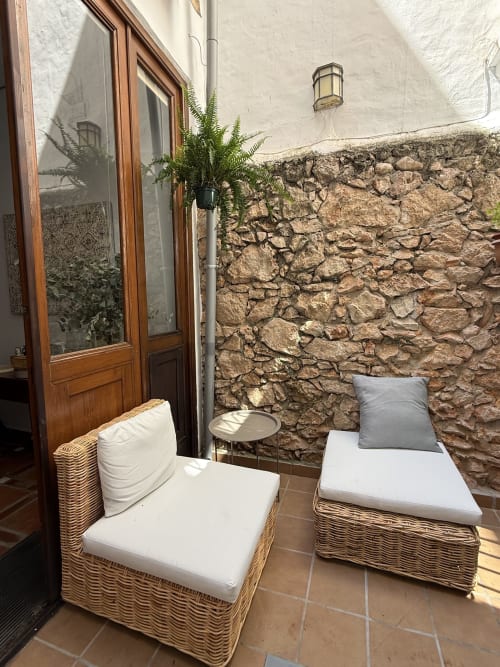 Acogedora zona de descanso en Hotelito Oliva con sillas de mimbre, muros de piedra y plantas en macetas junto a puertas de madera.