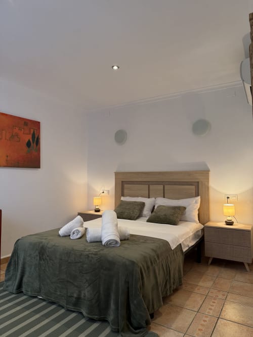 Moderna habitación doble en Hotelito Oliva con dos camas, cabecera de madera, iluminación cálida y altavoces blancos de techo.