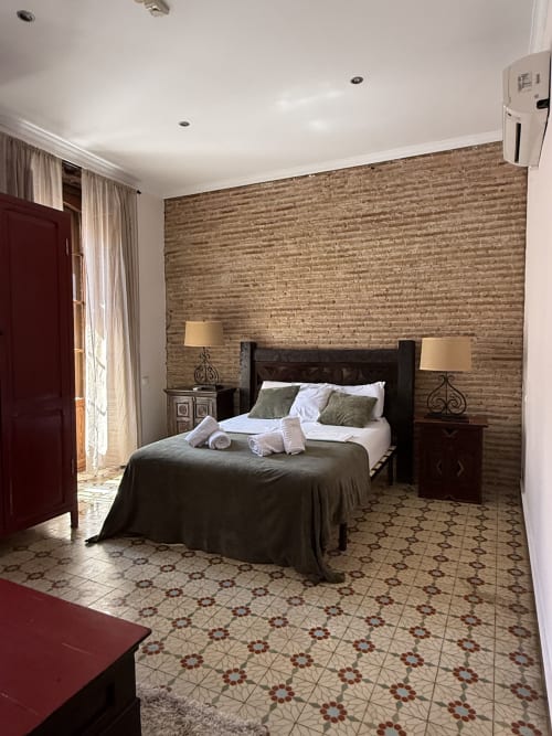 Amplia habitación doble en Hotelito Oliva con pared de ladrillo, cama de madera oscura, lámparas gemelas y piso de azulejos estampados