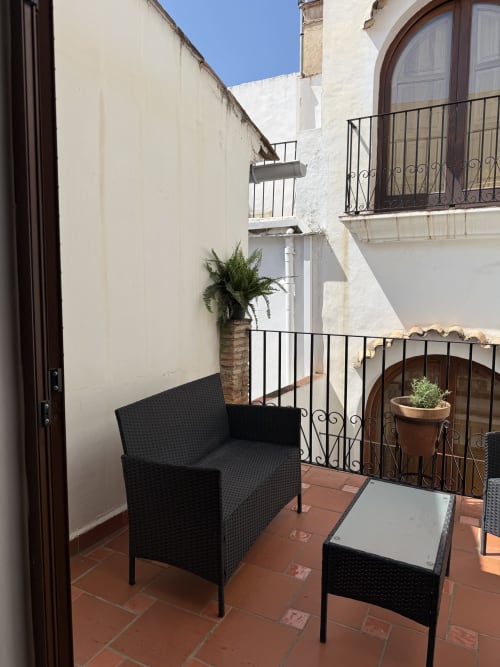 Acogedor rincón de descanso en el balcón del Hotelito Oliva Pensión en Oliva, España con silla de mimbre negra, mesa y plantas en macetas