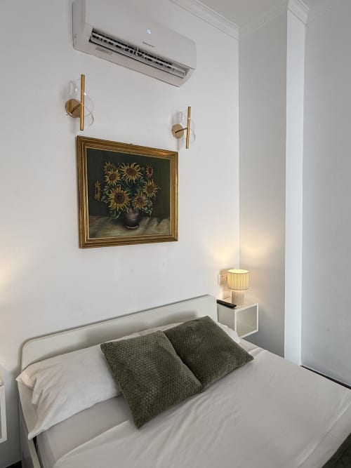 Dormitorio moderno en Hotelito Oliva con cama blanca, almohadas verdes, cuadro de girasoles, apliques de latón y aire acondicionado